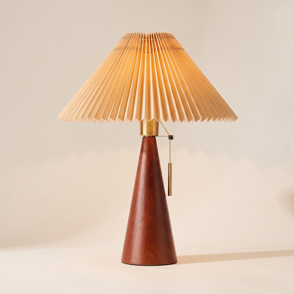 Walnut wood pleated table lamp with a beige pleated shade and a brass pull chain, switched on