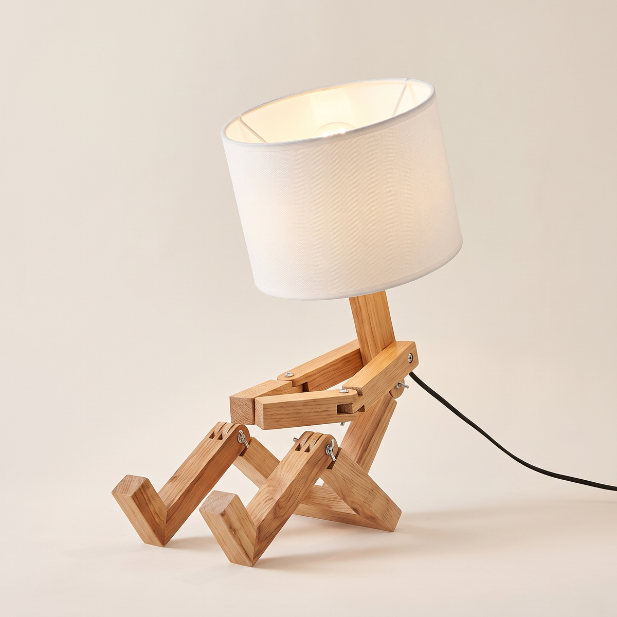 Jointed wooden robot desk lamp sitting down with a white fabric drum shade, switched on