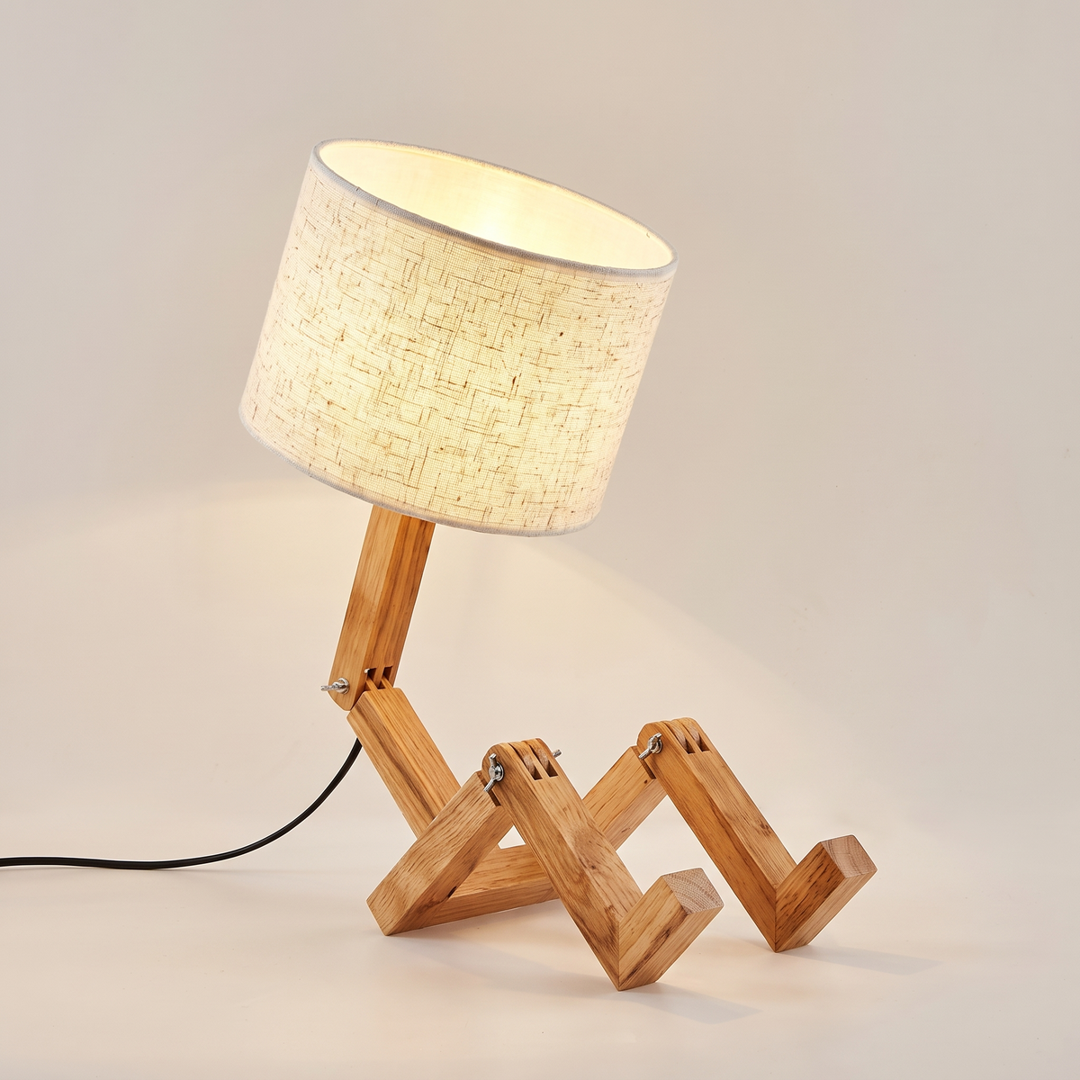 Jointed wooden robot desk lamp with a beige linen drum shade, seated and switched on