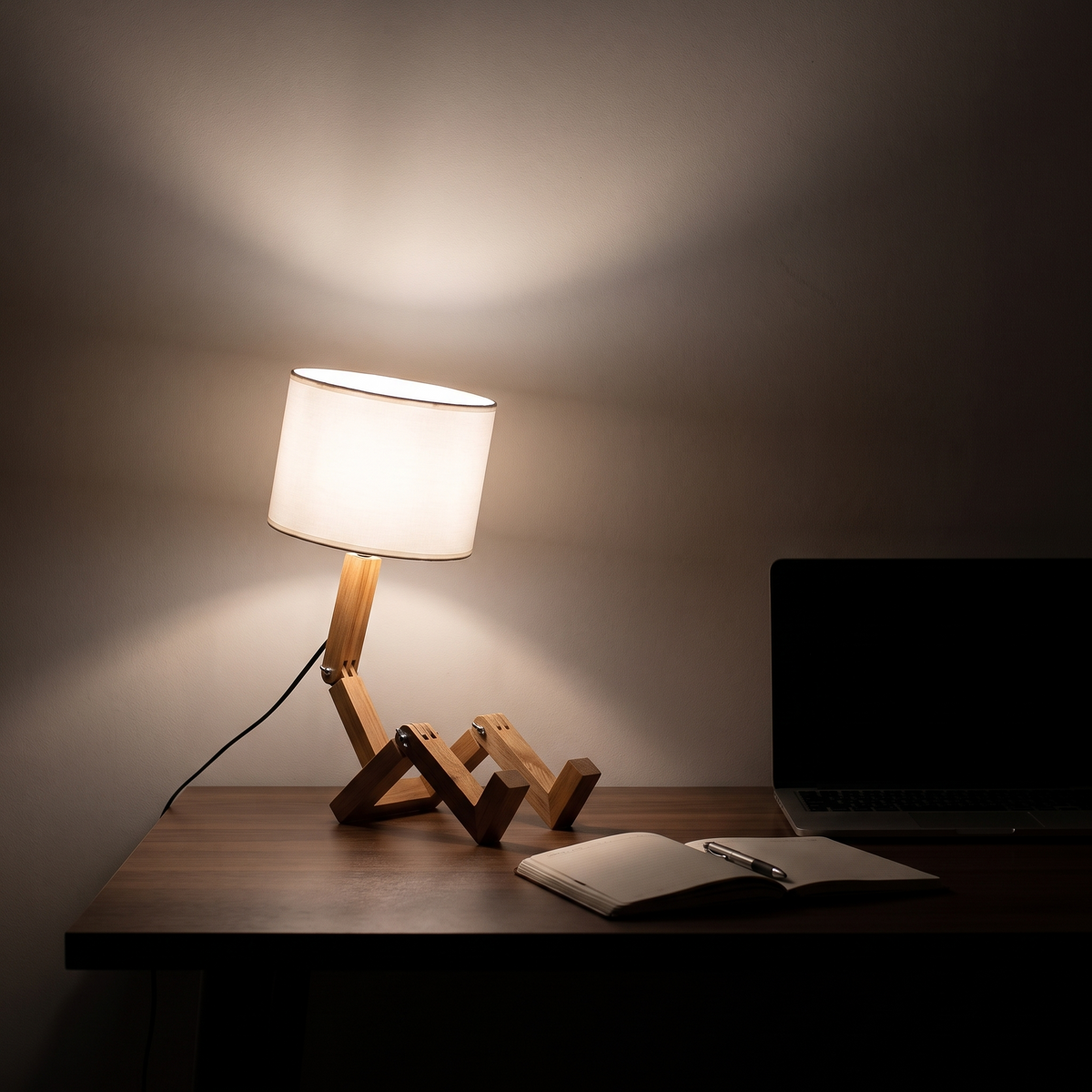 Jointed wooden robot desk lamp lit at night on a desk beside an open notebook