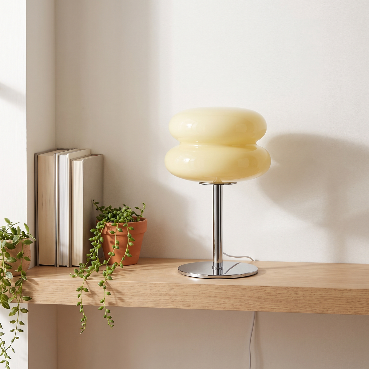 Beige macaron glass table lamp switched off on an oak shelf beside books and a plant