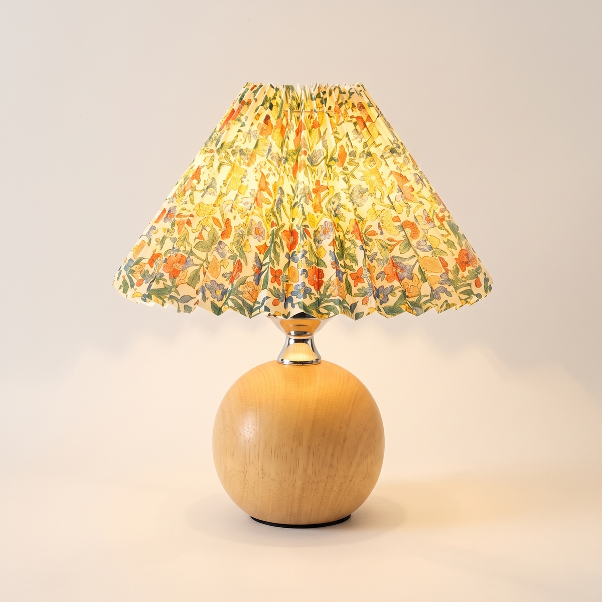 Light wood ball table lamp with a yellow and green floral pleated shade, switched on